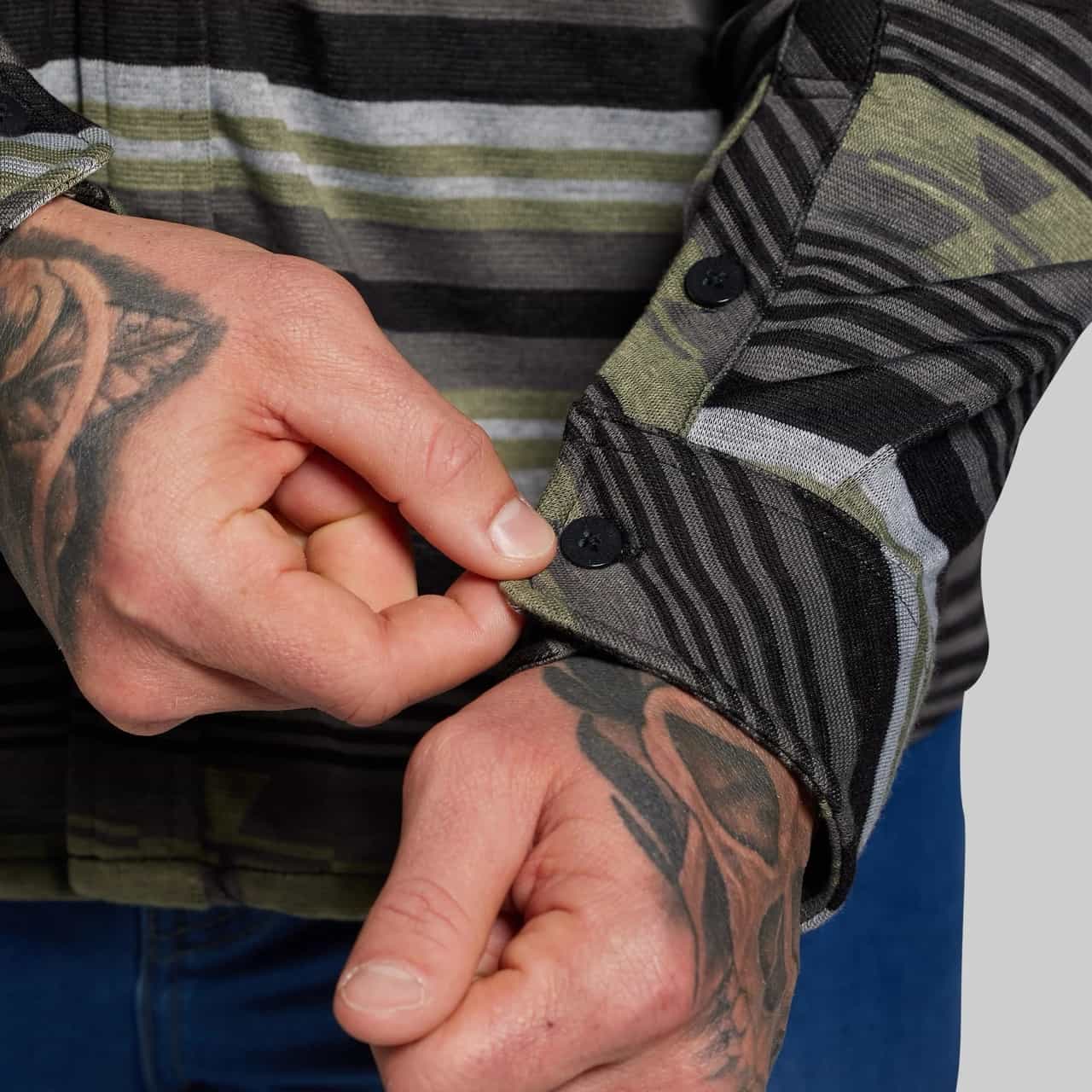 Person buttoning a jacket with tattoos on their arms, wearing a striped shirt.