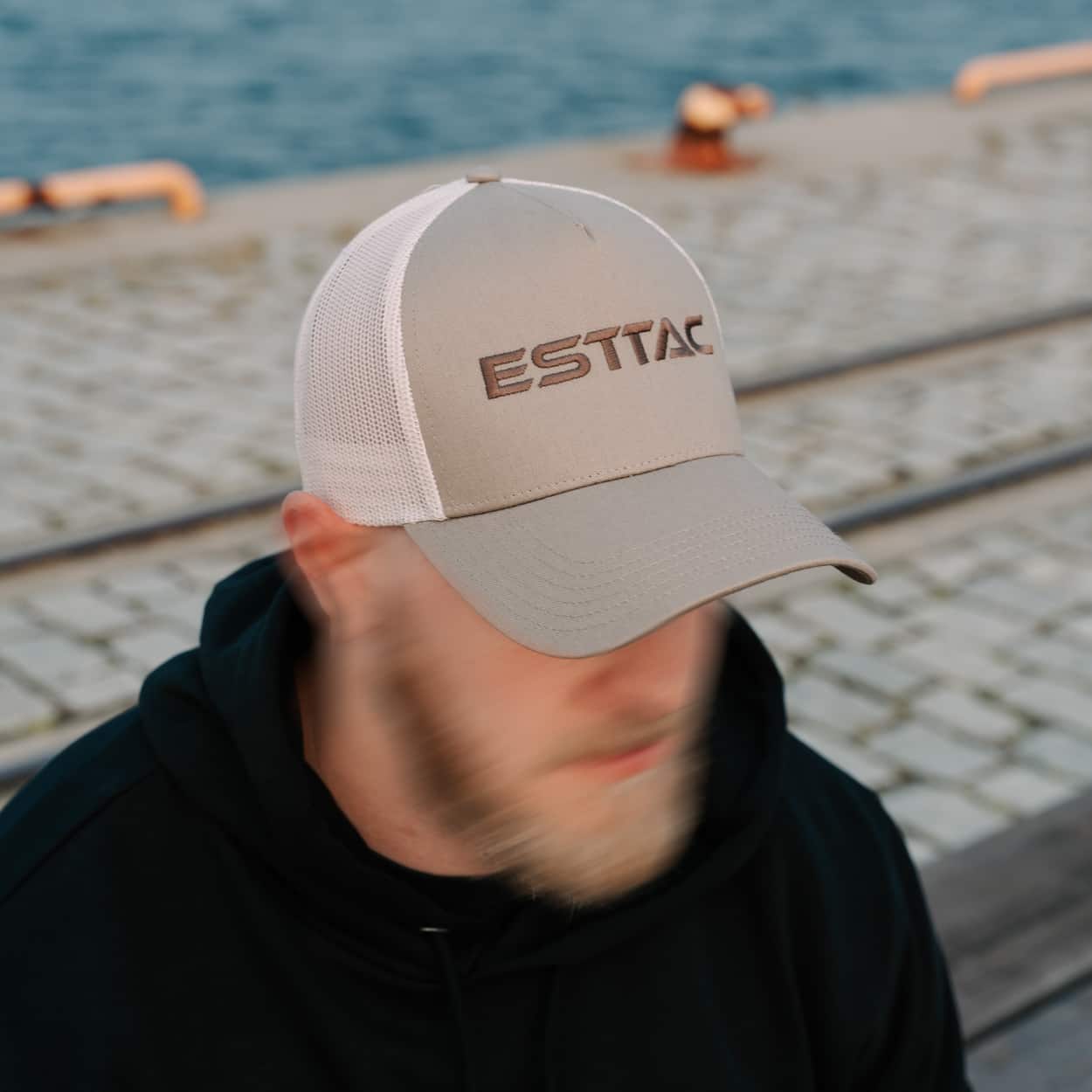 Person wearing a beige cap with 'ESTTAC' logo by a waterfront