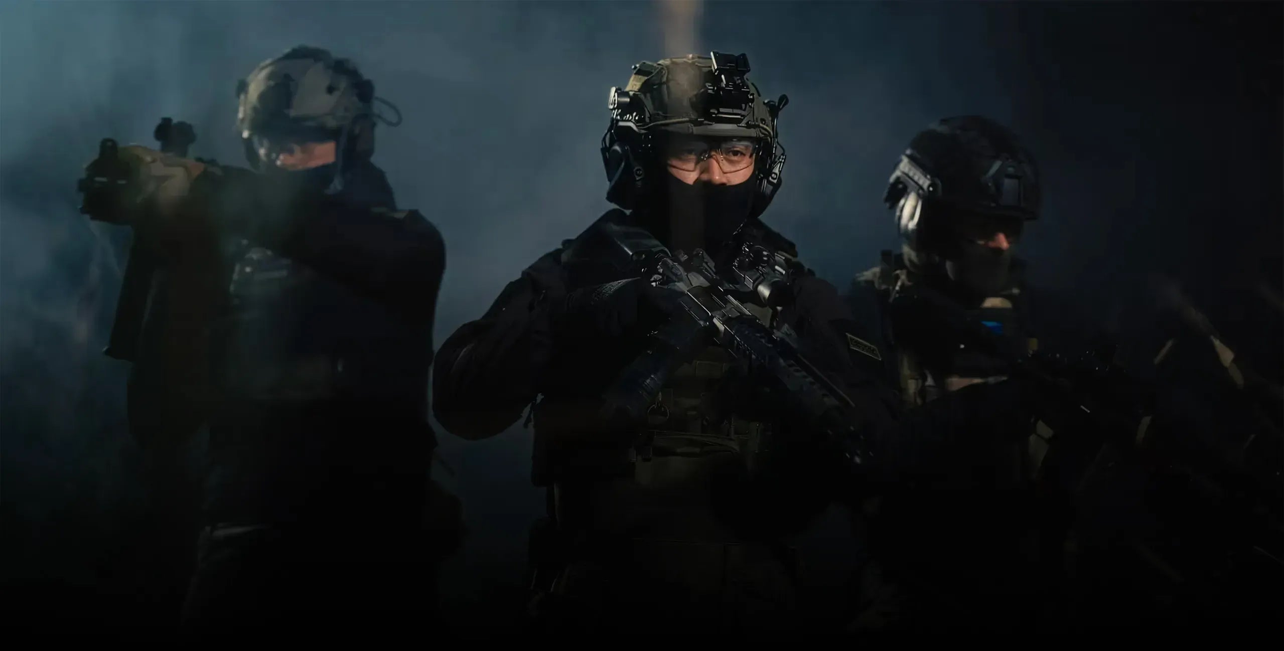 Three tactical soldiers in dark uniforms and helmets with night vision gear, standing in a smoky dark environment. The central soldier is in focus, while the two flanking soldiers are blurred in the background.