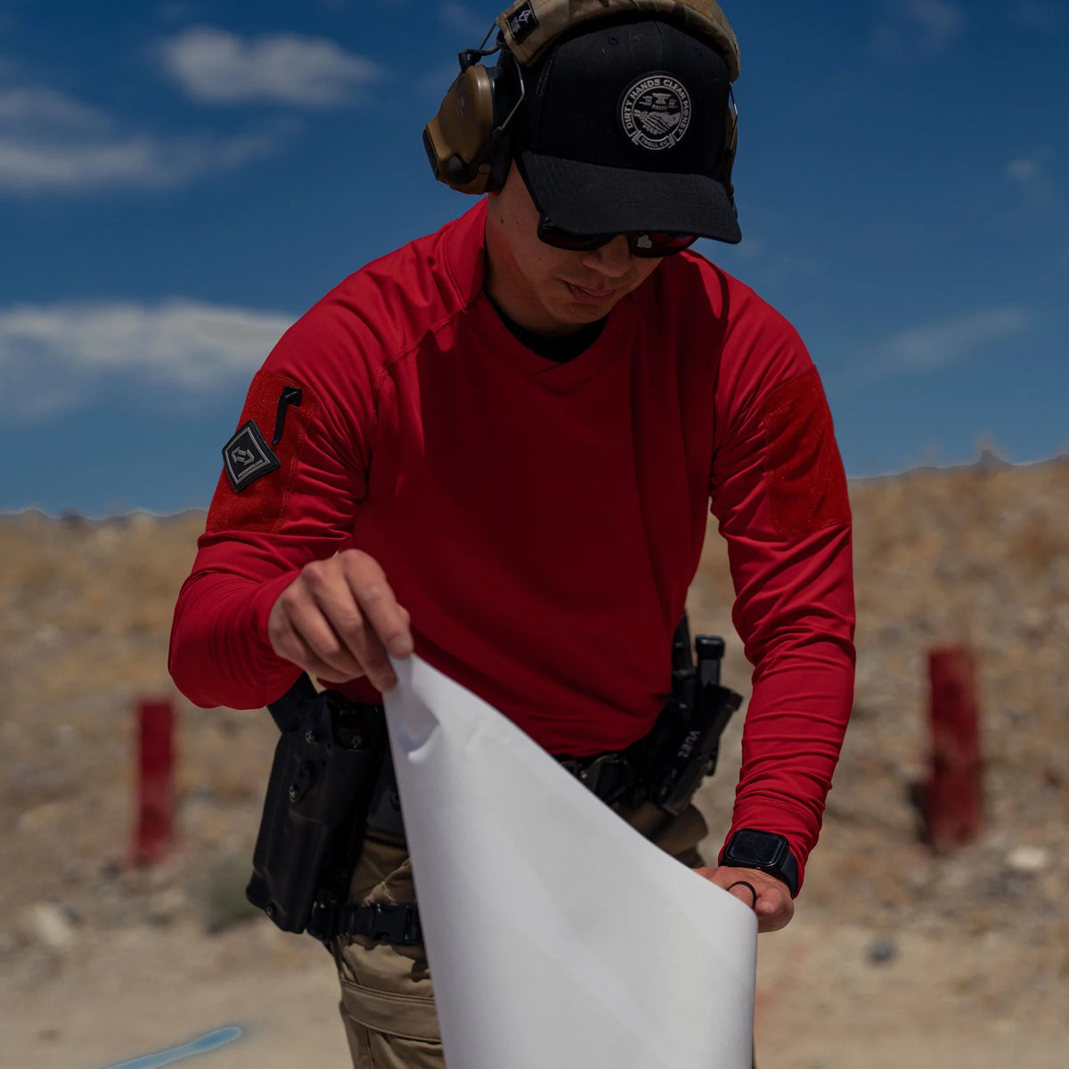 Vertx Men's Enduro Training Long Sleeve Shirt in red at an outdoor shooting range, front view while handling a target