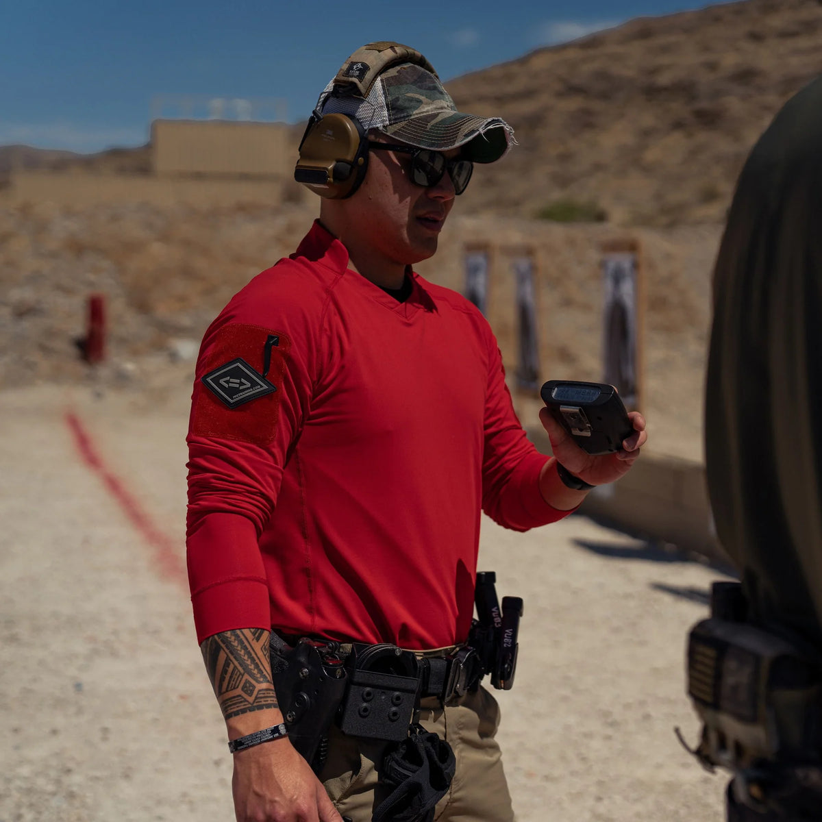 Vertx Men's Enduro Training Long Sleeve Shirt in red at an outdoor shooting range, front view with sleeve patch