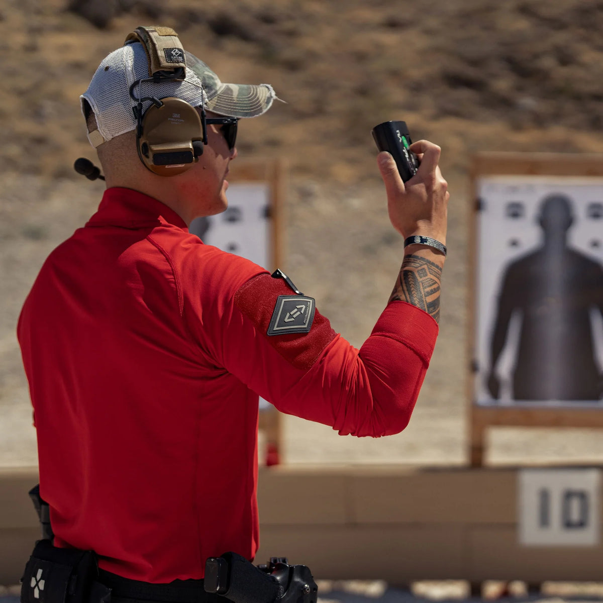 Vertx Men's Enduro Training Long Sleeve Shirt in red at an outdoor shooting range, rear side view with sleeve patch